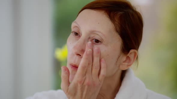 Headshot of Confident Mature Woman Applying Moisturizing Cream on Face Looking Away