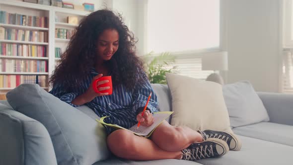 Cute Indian Teenage Girl Sits Comfortably on Sofa with Copybook in Living Room