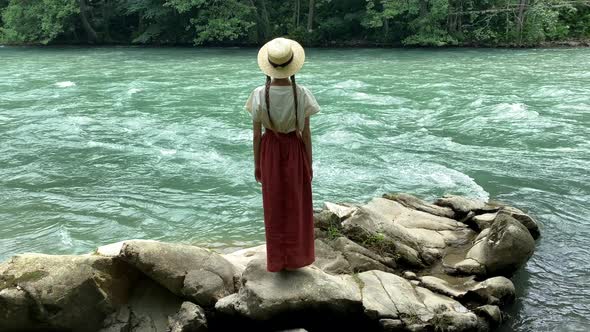 Young vintage woman in dress and straw boater hat stand near rippling mountain river, back view