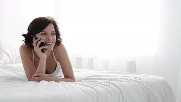 Woman Talking On Mobile Phone And Lying On Bed With White Linens