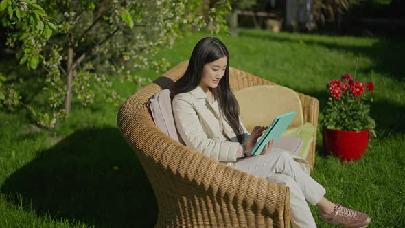 Young Slim Charming Asian Woman Smiling Surfing Internet on Tablet Sitting on Couch in Sunny Spring