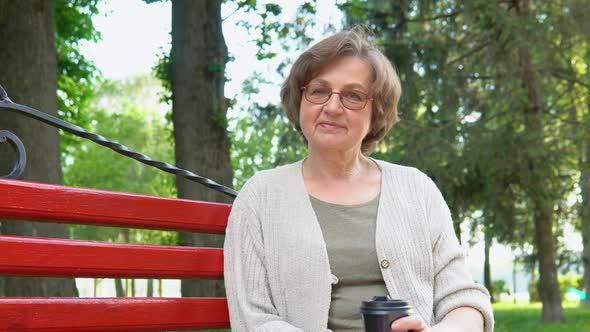 Portrait of Elderly Woman with Glasses and Coffee Smiling on Bench in Public City Park