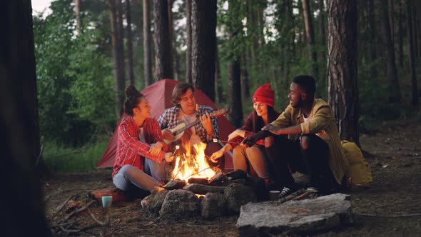 Relaxed Men and Women Travelers Are Singing Songs Around Campfire in Forest, Playing the Guitar and