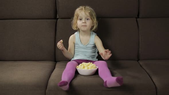 Girl Sitting on Sofa and Eating Corn Puffs. Child Watching Tv, Taste Puffcorns