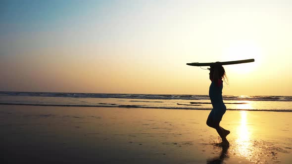 Little girl is running along the ocean sand beach with surfboard at sunset.