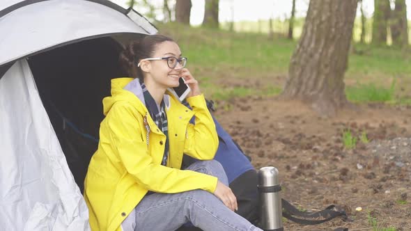 Young woman tourist sitting near a tent and talking on smart phone in forest.