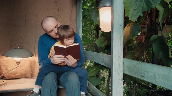 Man Reads a Paper Book for Small Child Sitting on the Porch of a Village House