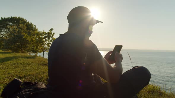 Man Tourist is Resting on the Seashore with a Smartphone in His Hand