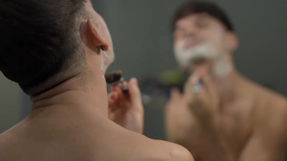 Back View Young Man Shaving Chin with Blurred Reflection in Mirror at Background