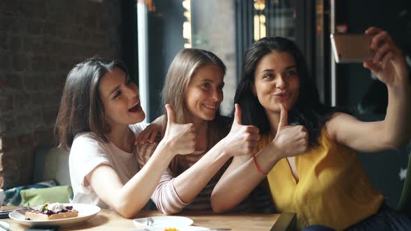 Happy Young Ladies Taking Selfie with Thumbs-up Gesture in Cafe Laughing