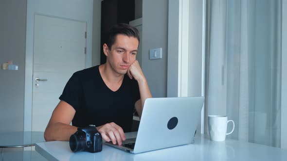 Young Photographer Using Laptop Processing Images on His Laptop and Drinking Coffee at Home