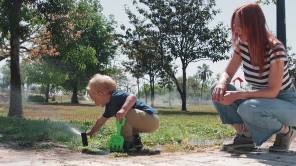 Little Boy Playing with Water Sprinkler  His Mother Sitting Nearby