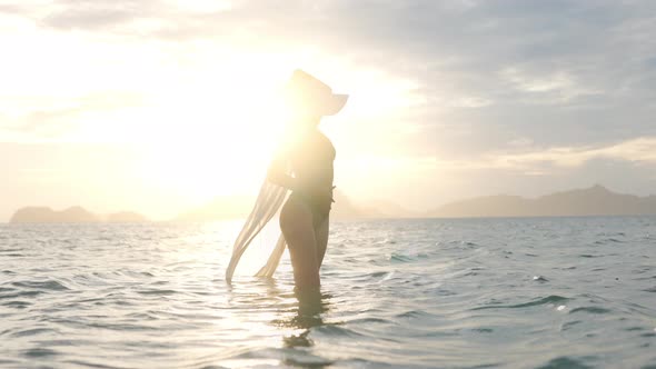 Woman In Bikini With Sun Hat Standing In Sea At Sunset