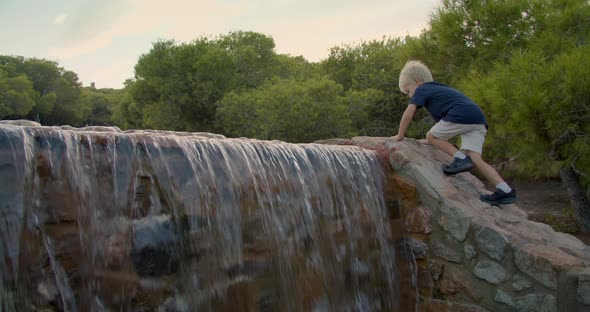 Cute Blond Boy Climbs the Mountain Near the Falling Water at Evening in Parkland