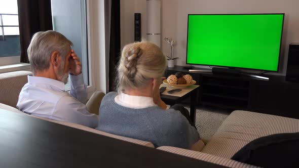 An Elderly Couple Sits on A Couch in A Living Room, Watches TV with A Green Screen and Acts Upset