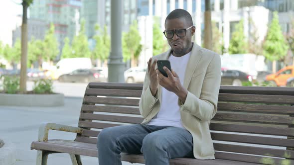 African Man Reacting to Loss on Smartphone While Sitting Outdoor on Bench