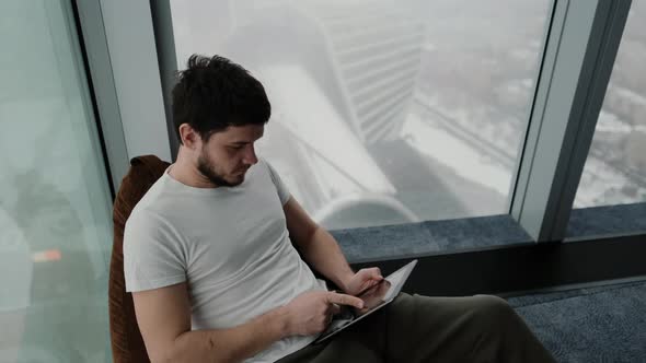 Gorgeous Man Sitting on Pouf Using Tablet in Modern Apartment Texting Scrolling Through Technologies