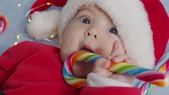 Smiling Funny Newborn Baby in Santa Claus Hat Red Bodysuit Play on White Soft Bed Lick Sweet