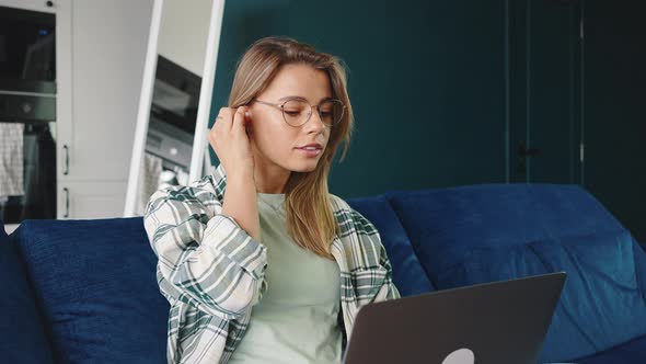 Young Blonde Woman Putting on Wireless Earphones and Working on Laptop Sitting Cozily on Sofa Ta