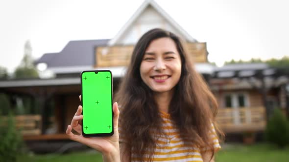 An Asian Girl Shows a Phone with a Green Screen Against the Background of Her New Home