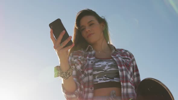 Smiling caucasian woman using smartphone and holding skateboard at a skatepark