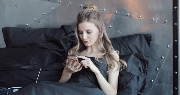 Gorgeous Blondie Checking Her Mobile Phone Sitting in Bed Under Black Blanket