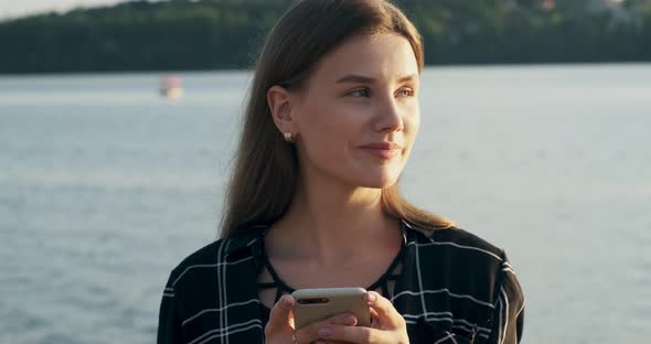 Close Up Portrait of Young Attractive Woman Smiling and Looking at Smartphone Near Beach at Sunset