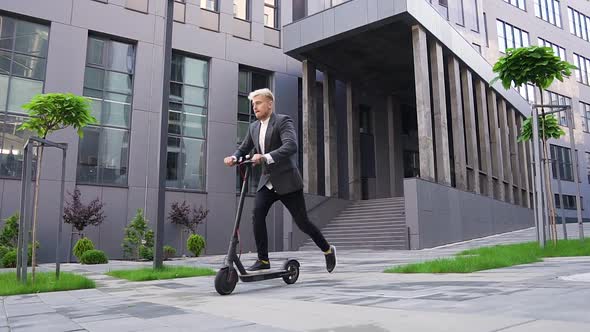 Young Light-Haired Guy Enjoying His Ride on Own E-Scooter Near Beautiful Architectural Building