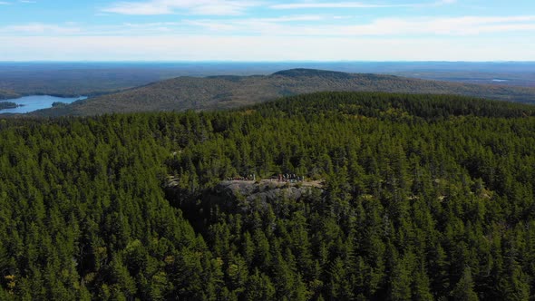 Aerial drone shot pulling back over a large green forest with lakes and a rocky outcrop in the Maine