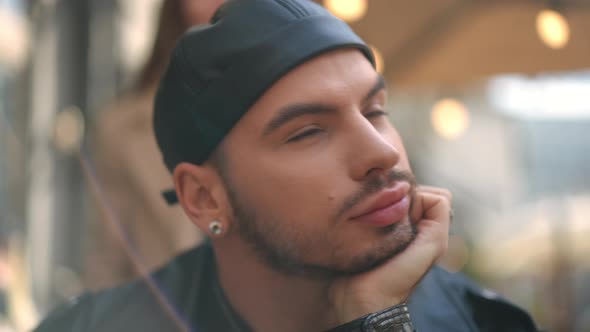 Headshot Portrait of Thoughtful Young LGBTQ Man Sitting in Sidewalk Cafe As Woman Walking at