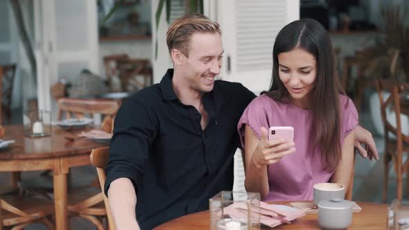 Young Man and Woman Sitting in Cafe, Looking at Smartphone, Laughing and Talking