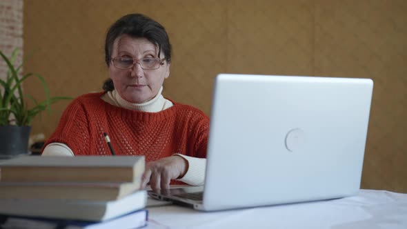 Modern Elderly Woman Writes Information in a Notebook While Studying Online Using Modern Technology