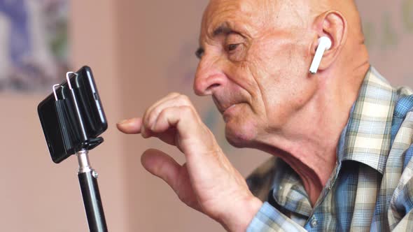 Caucasian pensioner 70-75 years old in headphones at home holds a tripod with a smartphone and click