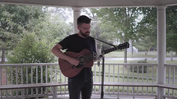 Man playing guitar and singing under a gazebo in the rain - Wide rotation around the front shot