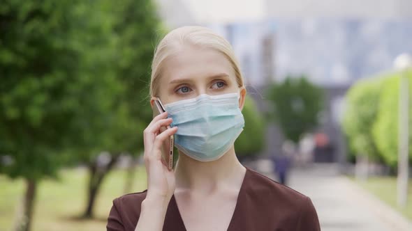 A Young Woman in a Medical Mask is Talking on the Phone on the Street Against the Backdrop of a Big