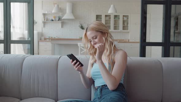 Happy Blonde Woman Using Smartphone Sitting on the Couch in Cozy Stylish Living Room Chatting and