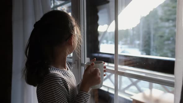 Happy Little Girl with a Cup of Tea Near the Window Looking Outside the House