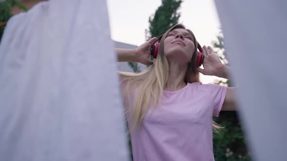 Young Happy Woman in Headphones Dancing to Music with White Clean Laundry Hanging on Rope at Front