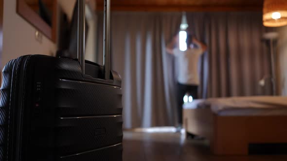 Closeup Black Travel Bag in Hotel Room with Blurred Young Man Opening Curtains at Background