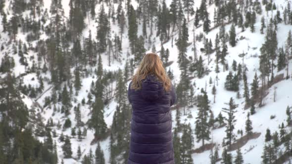 Blonde Girl Raises Her Hands Up Against the Background of Snowy Pine Forest in the Mountain