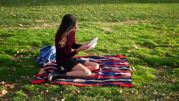 Beautiful young hispanic woman college student reading a book outdoors in the grass before class on