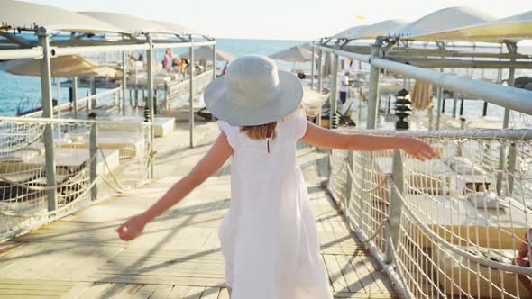 a Little Girl Blonde in a White Dress and a Widebrimmed Hat Runs on the Pier