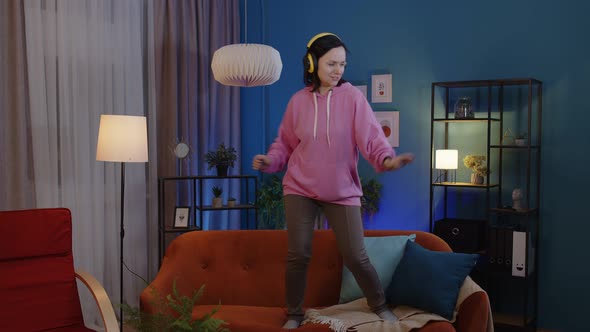 Happy Adult Woman in Headphones Dancing and Jumping While Listening Music at Home Alone on Sofa