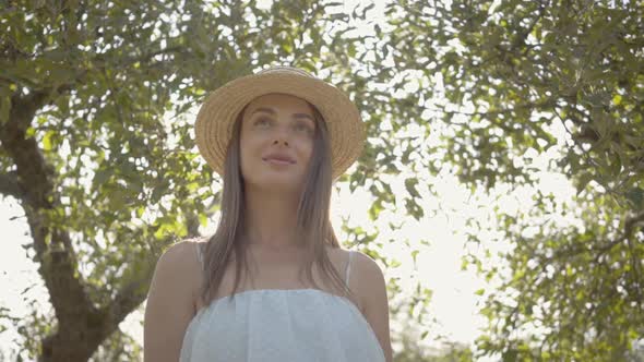 Attractive Young Woman in Straw Hat and Long White Dress Standing in the Green Summer Garden. Sun