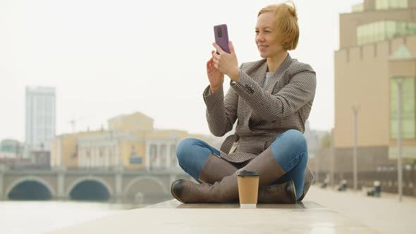 40Yearold Beautiful Woman Sits on the Embankment and Takes a Photo of the Cityscape with Her