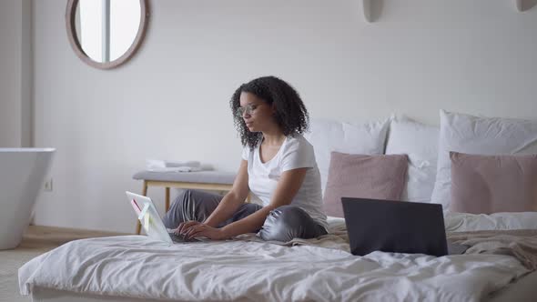 Wide Shot of Focused Beautiful African American Woman Sitting on Bed Typing on Laptop Keyboard