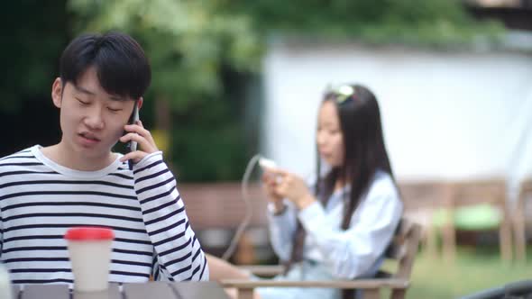 Asian Teen Boy Talking on Cell Phone at Cafe Table Outdoors