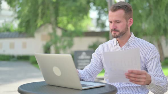 Middle Aged Man with Laptop Reading Documents in Outdoor Cafe