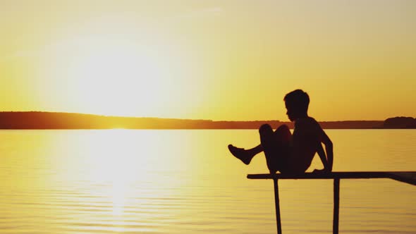 Little Boy is Sitting in a Lotus Position on the Edge of a Bridge Above a River