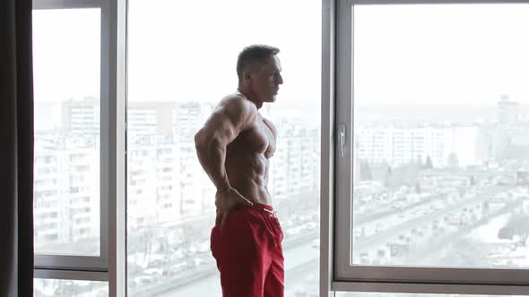 Muscular Shirtless Man Standing on the Background of Panoramic Windows and Warming Up His Waist By